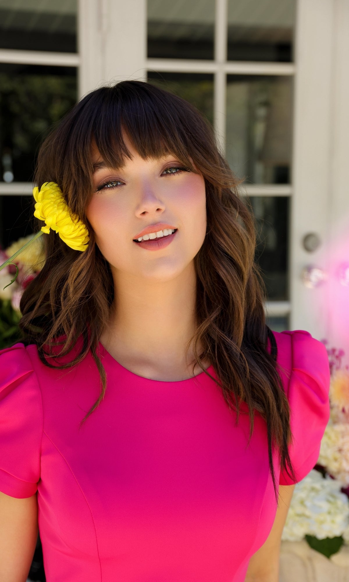 A woman with wavy brown hair smiles outdoors, wearing the bright pink Puff-Sleeve Homecoming Dress: Ava Presley 29801, with a yellow flower tucked near her face.
